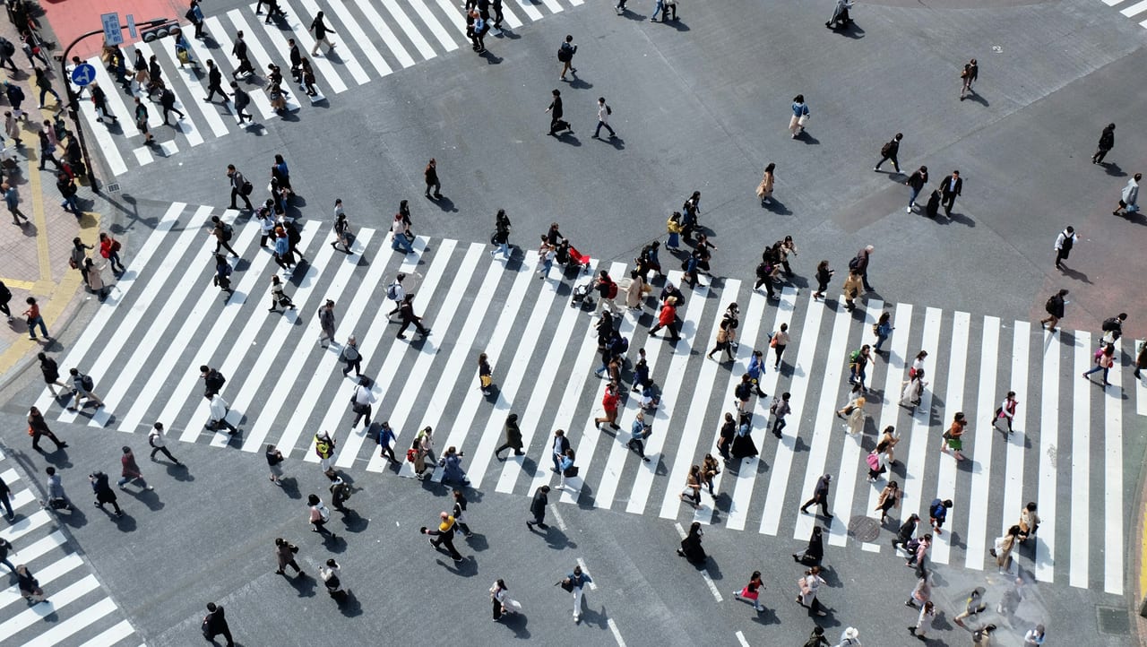 Crowds stream in every direction across the painted lanes of the Shibuya scramble crossing, seen from above in daylight.