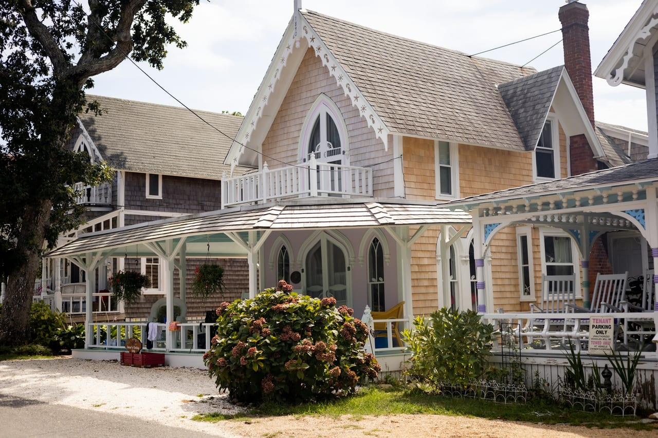A white Victorian-style house with a porch stands beside a tree-lined street.