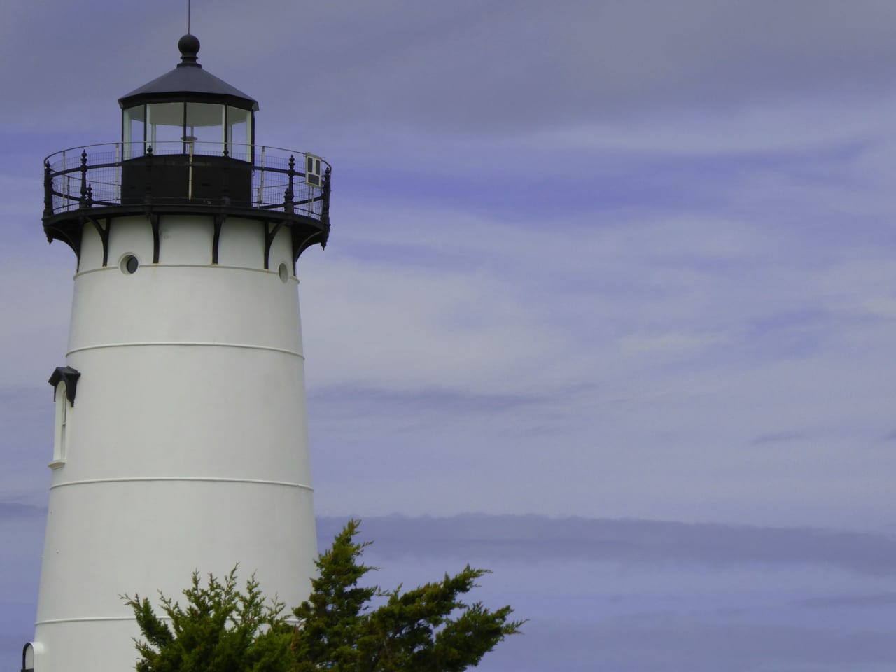A white lighthouse with a black lantern room stands against an overcast sky, with tree tops visible at the base.