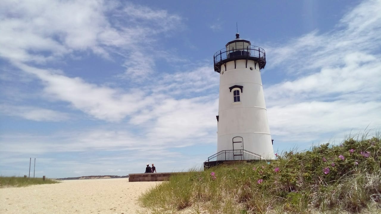 A white lighthouse stands beside a sandy beach under a blue sky.