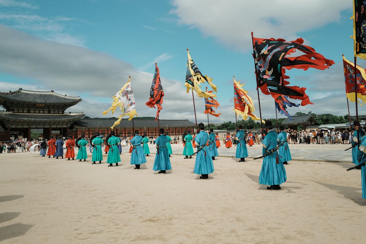 People in colorful traditional clothing stand beneath flags in a Seoul palace courtyard.