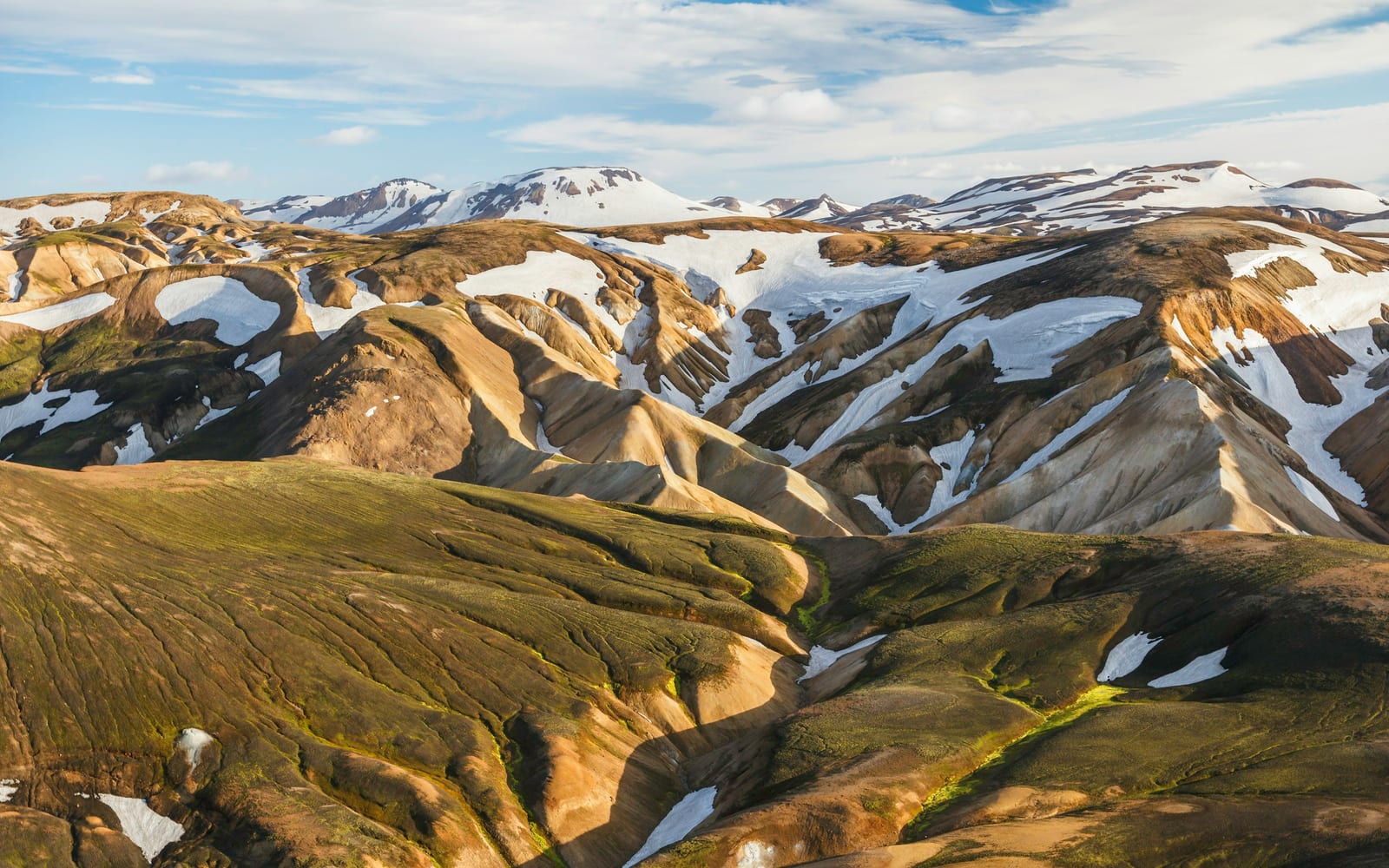 A broad view of colorful, snow-streaked mountains along the Laugavegur trail under a partly cloudy sky.