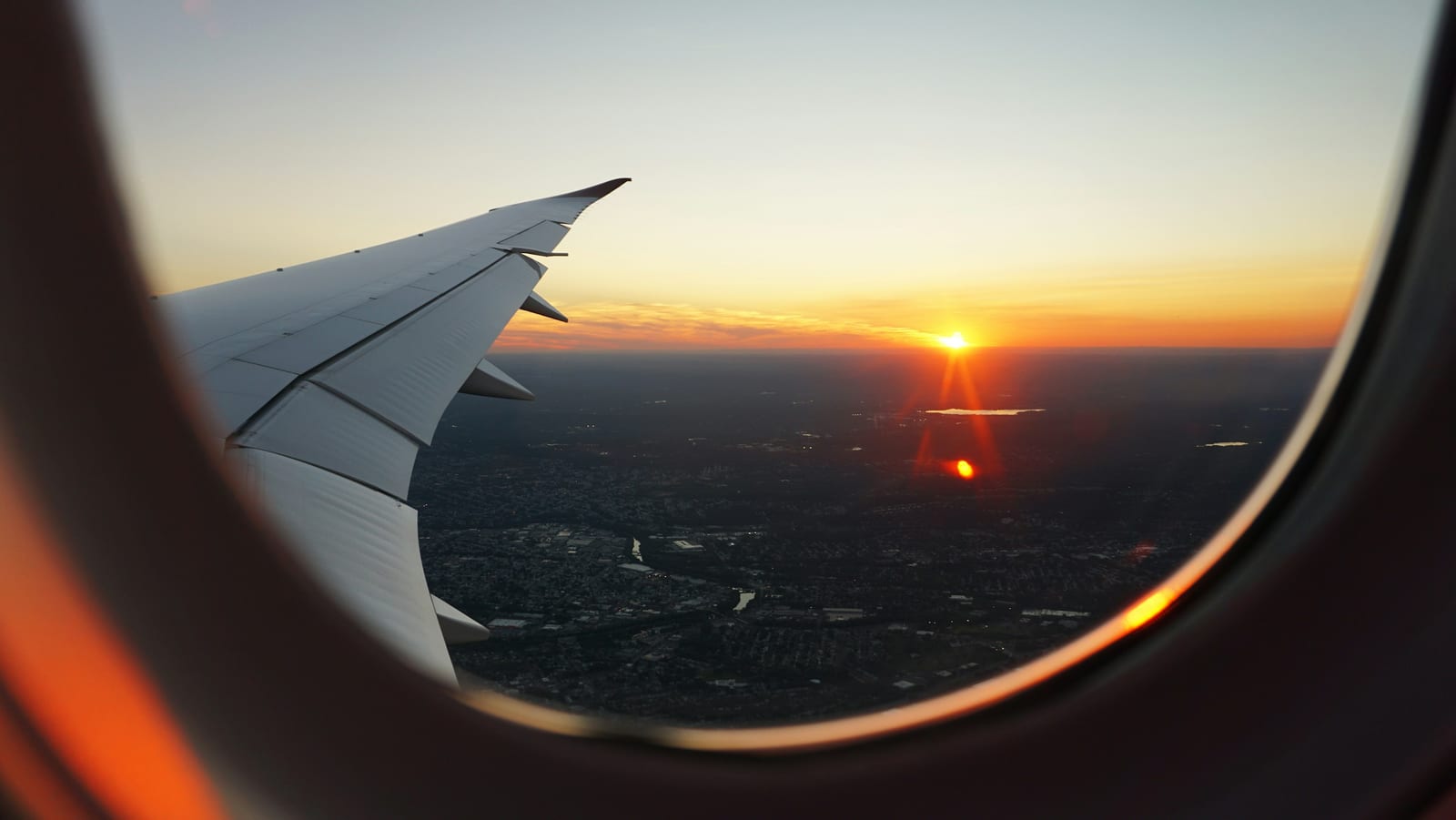 An airplane wing framed by a plane window against an orange sunset sky.