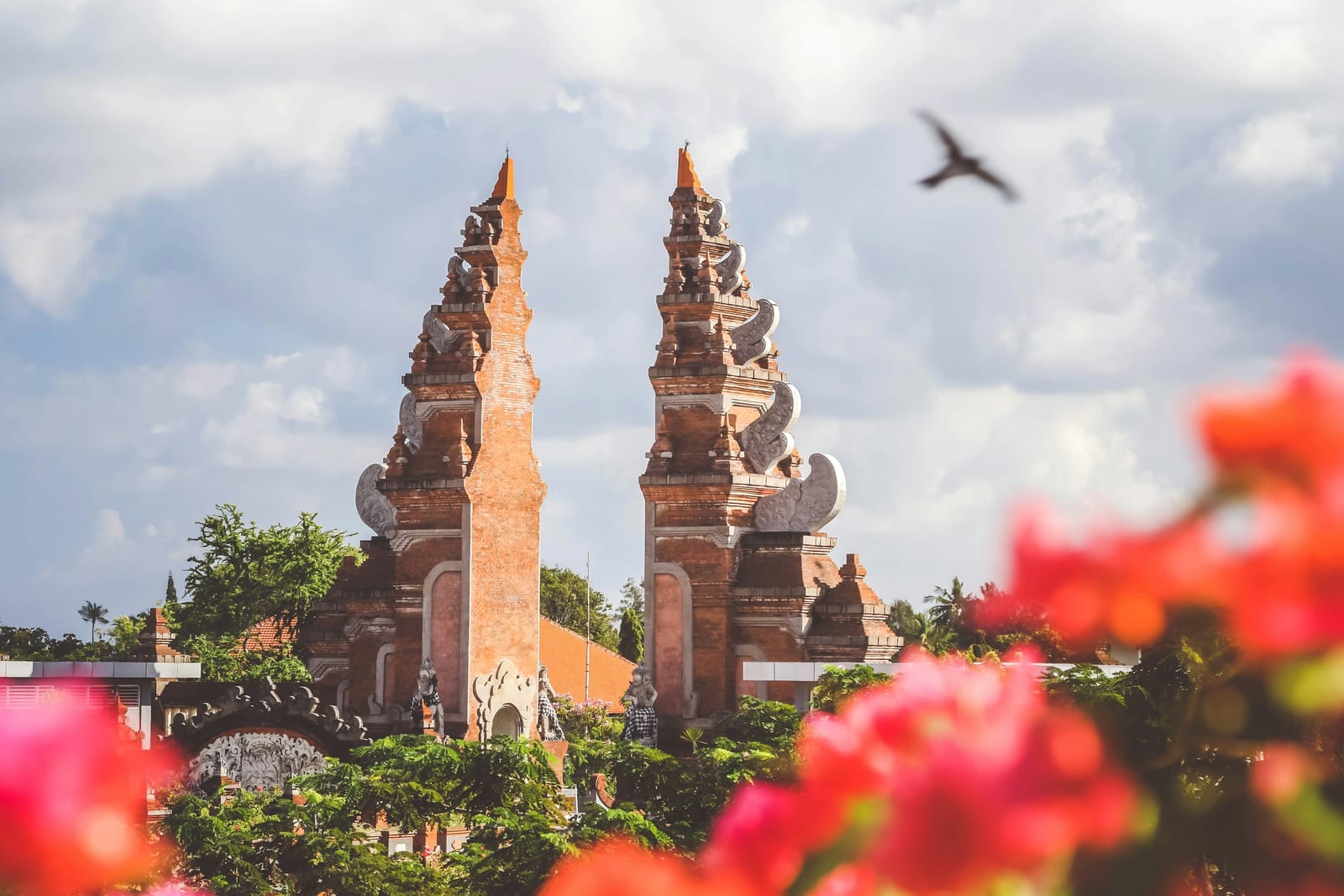 A Balinese split temple gate rises behind bright red tropical flowers under a partly cloudy sky.