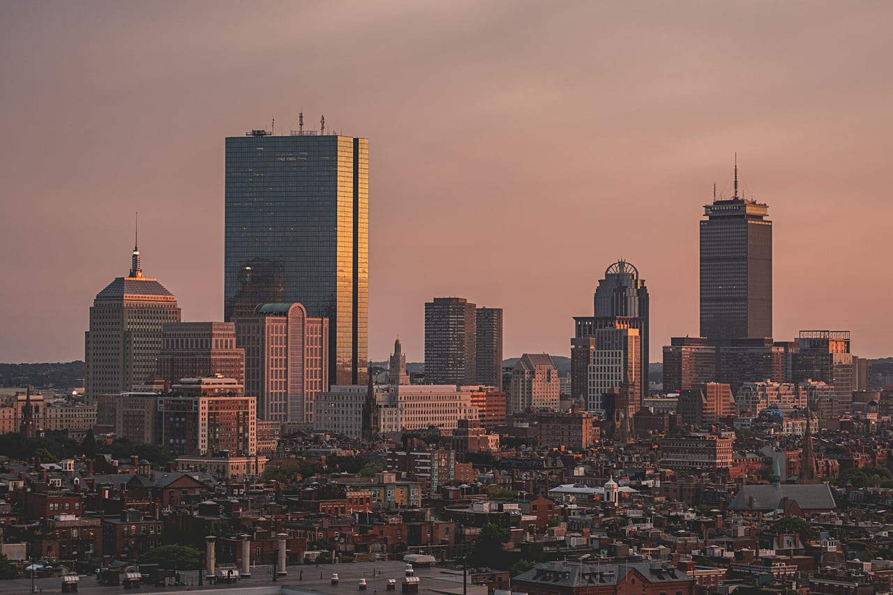The Boston skyline spreads out at sunset from a high observation point, the Charles River winding through the city.