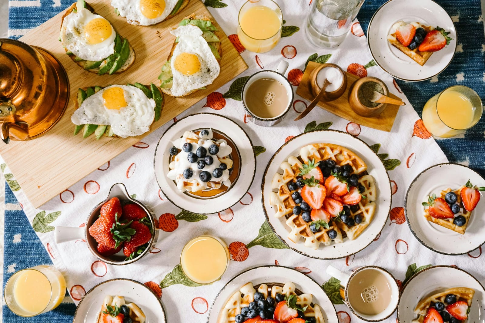 An overhead breakfast spread includes waffles with berries, eggs, coffee, and orange juice on a patterned tablecloth.