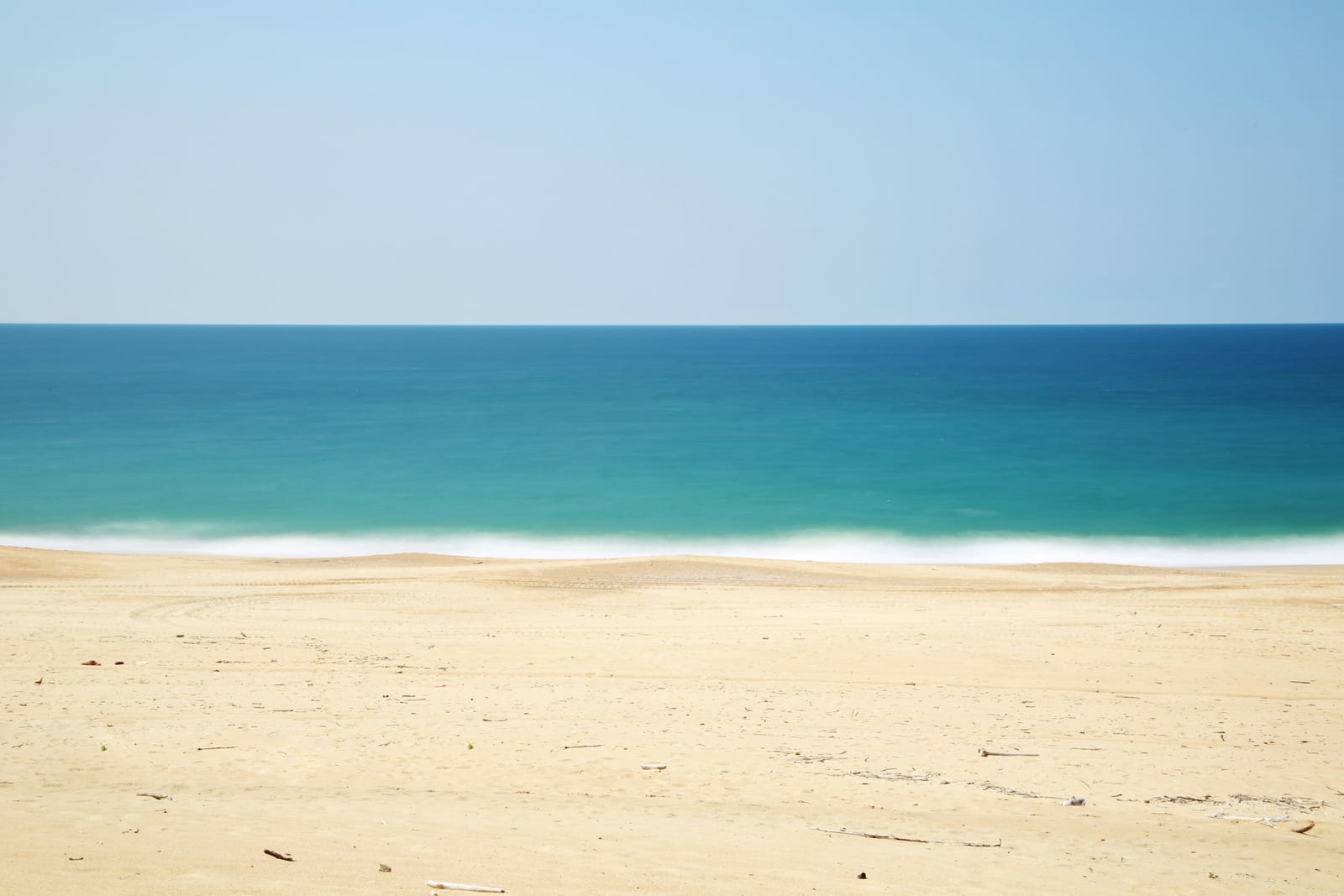 An empty sandy shoreline meets clear blue-green ocean water under a clear sky.