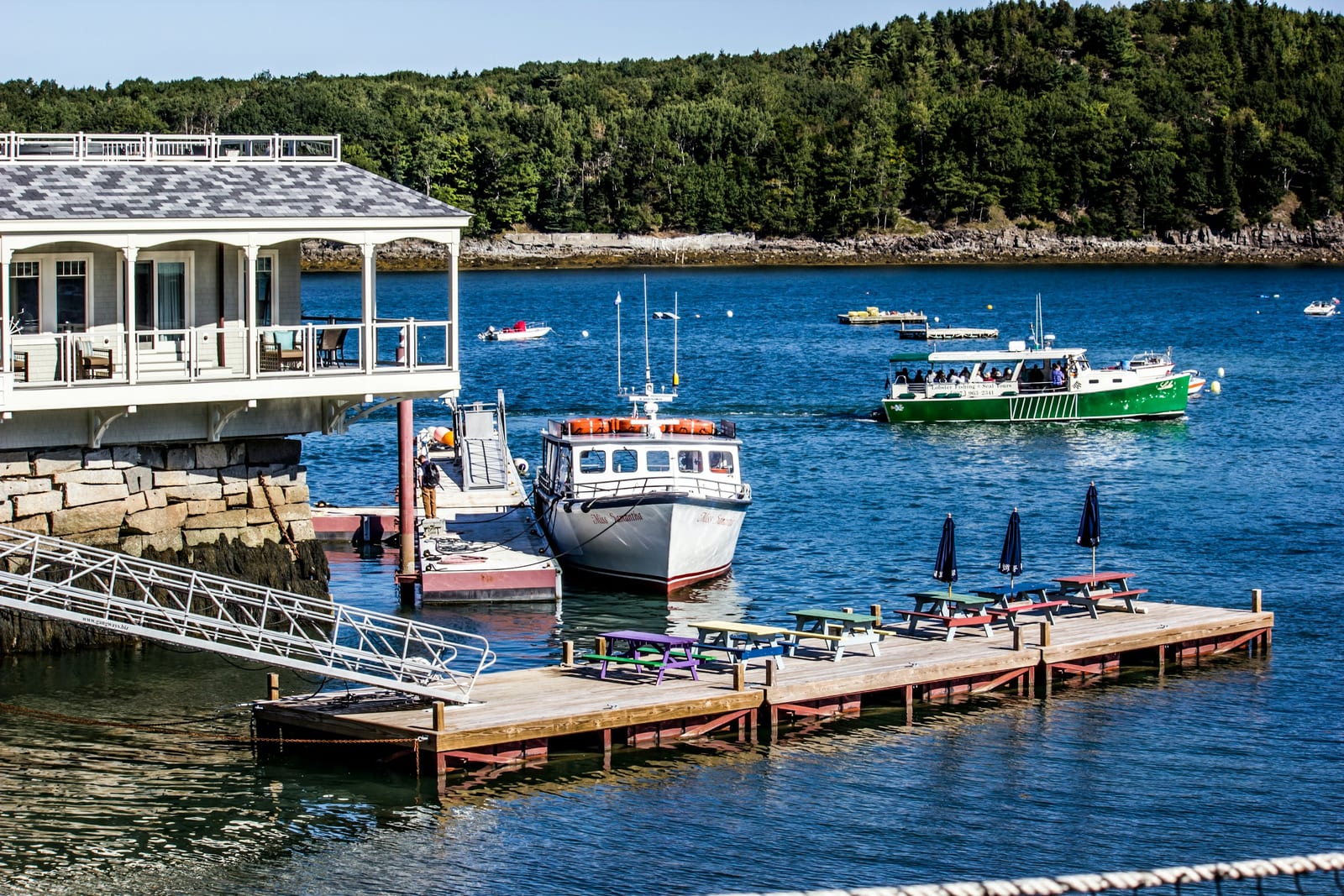 A harborside building with a wraparound porch overlooks a dock where several boats are moored on blue water, with a wooded shoreline behind.