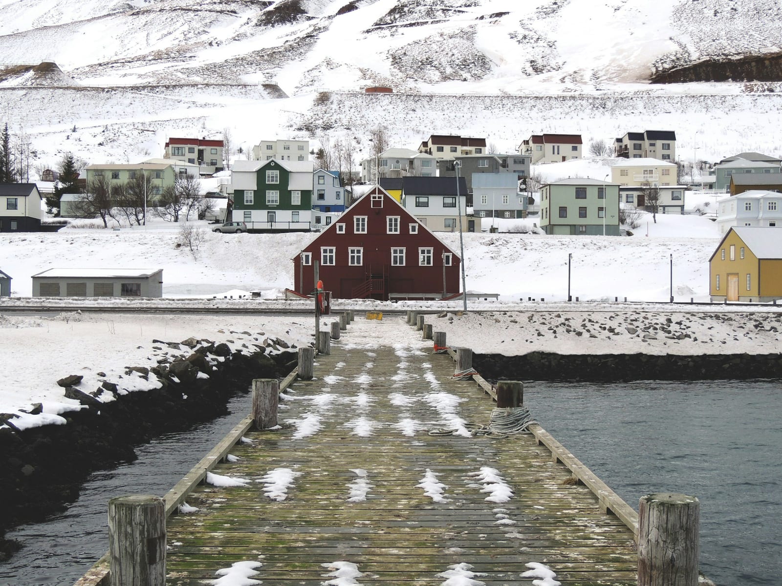A snow-dusted harbor pier leads toward a red waterfront building and a hillside town in Iceland.