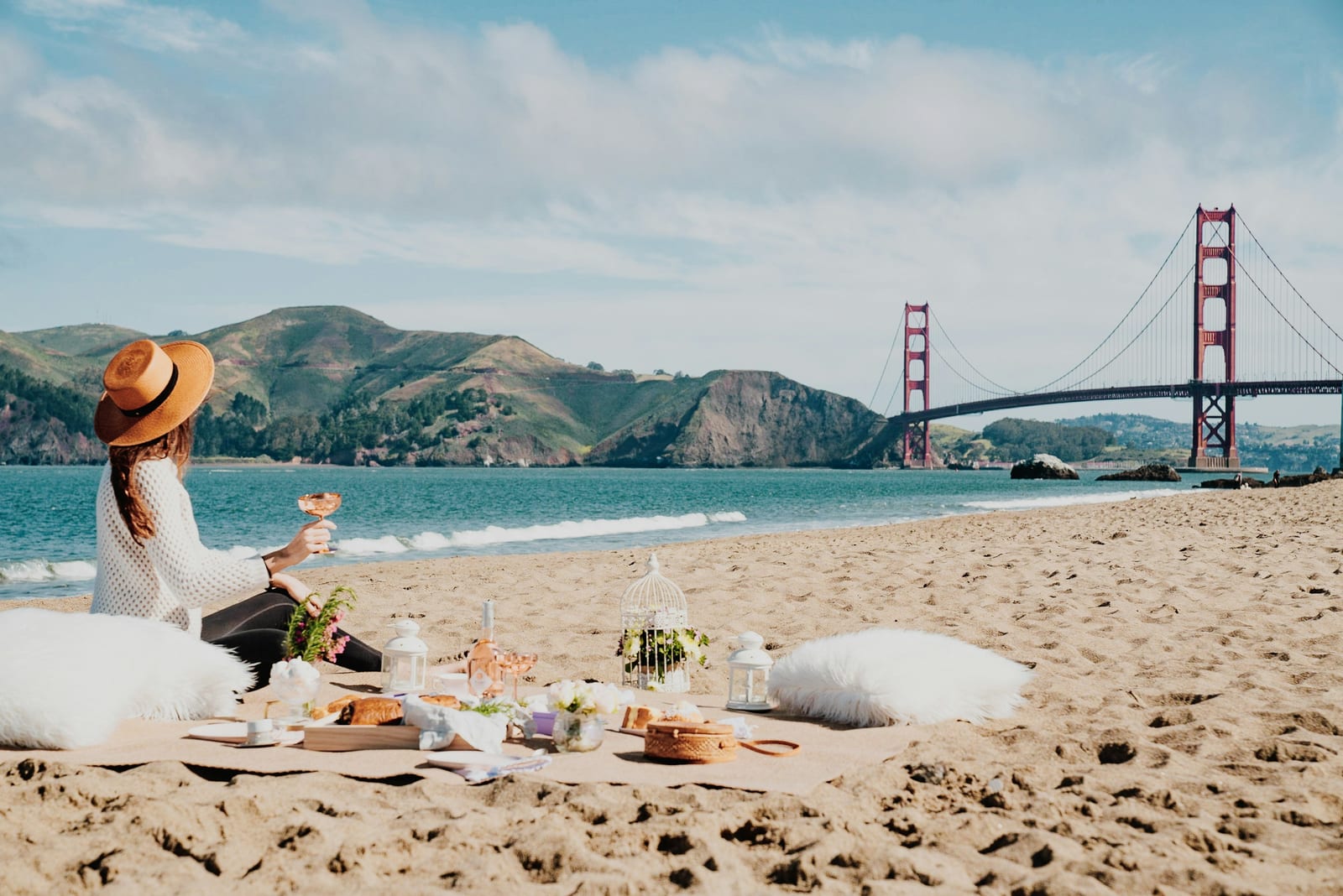A picnic spread with food and drinks sits on a sunny sandy beach beside a seated person, with a suspension bridge across the bay.