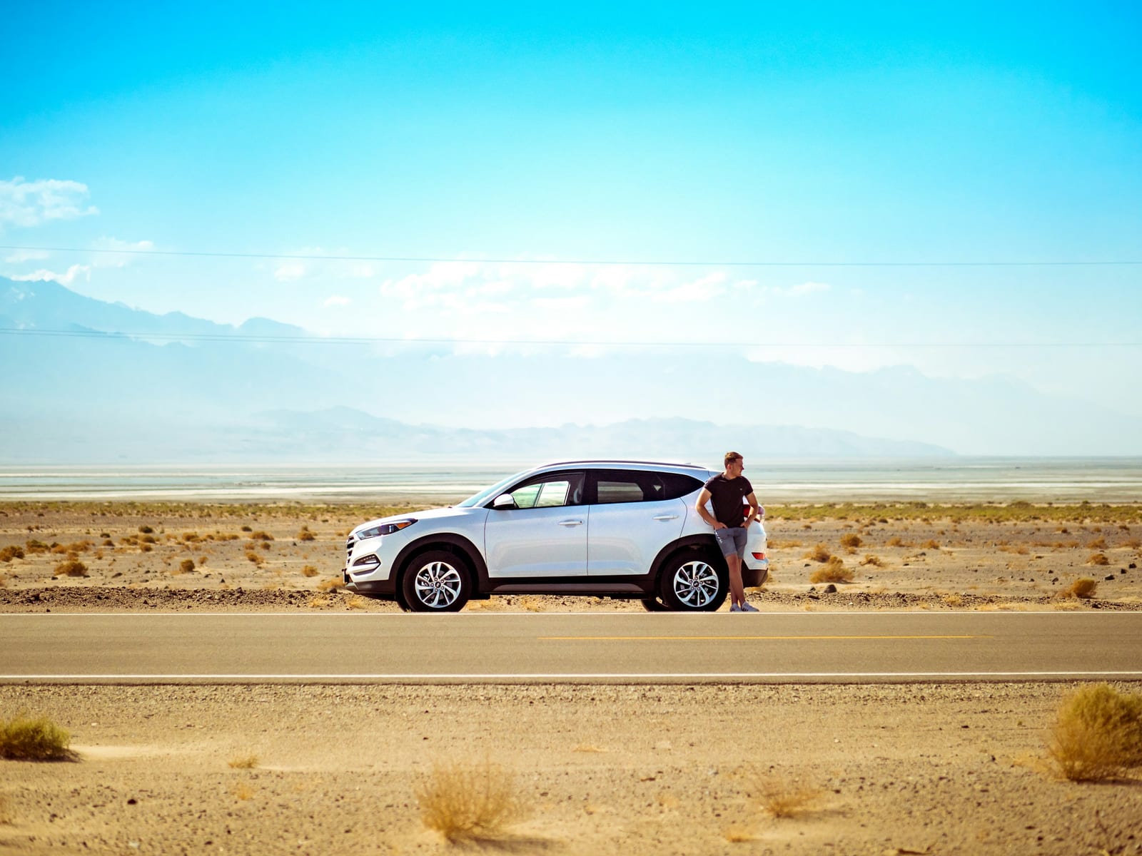 A white rental-style SUV is parked beside a sunlit road in a wide desert landscape.