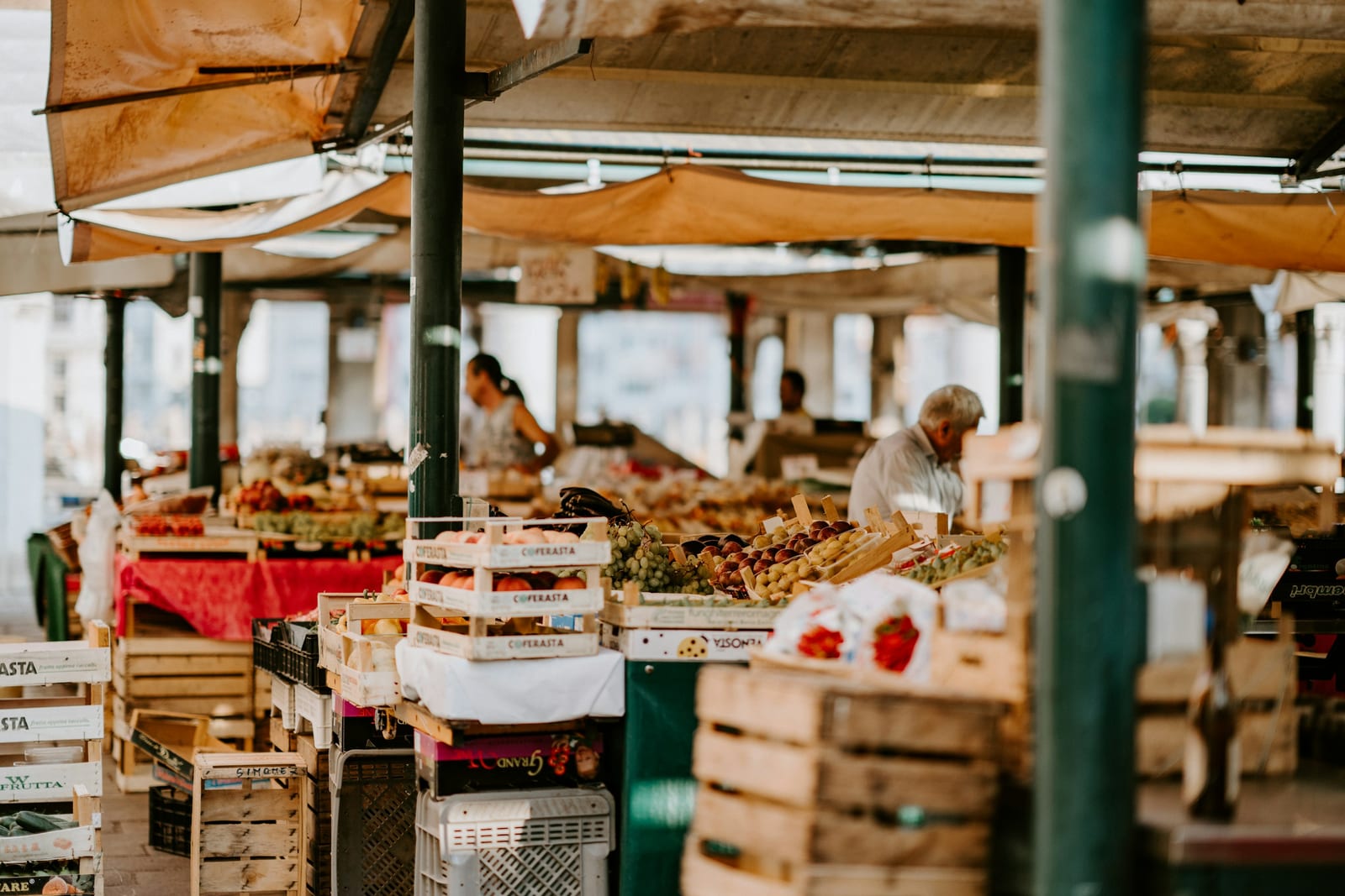 An open-air Roman food market with wooden crates of produce under awnings and a vendor at a stall.