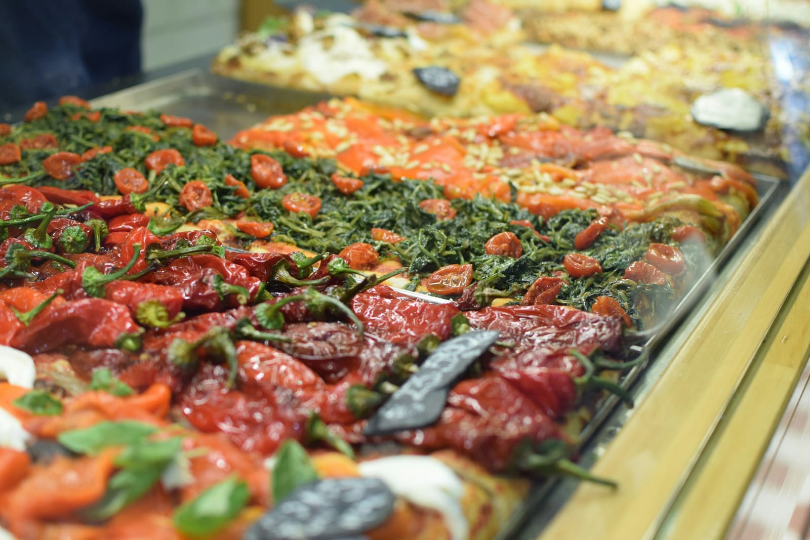 A display counter of Roman pizza al taglio topped with tomatoes, greens, and vegetables, sold by the slice.