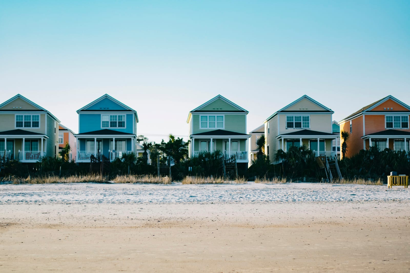 A row of colorful beach houses stands behind a sandy beach under a clear blue sky.