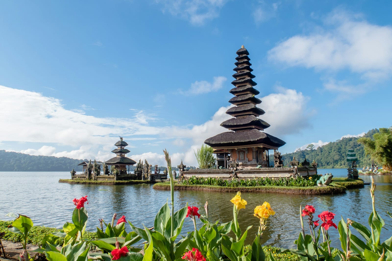 A Balinese temple with a tiered pagoda roof stands beside a lake, with flowers in the foreground.