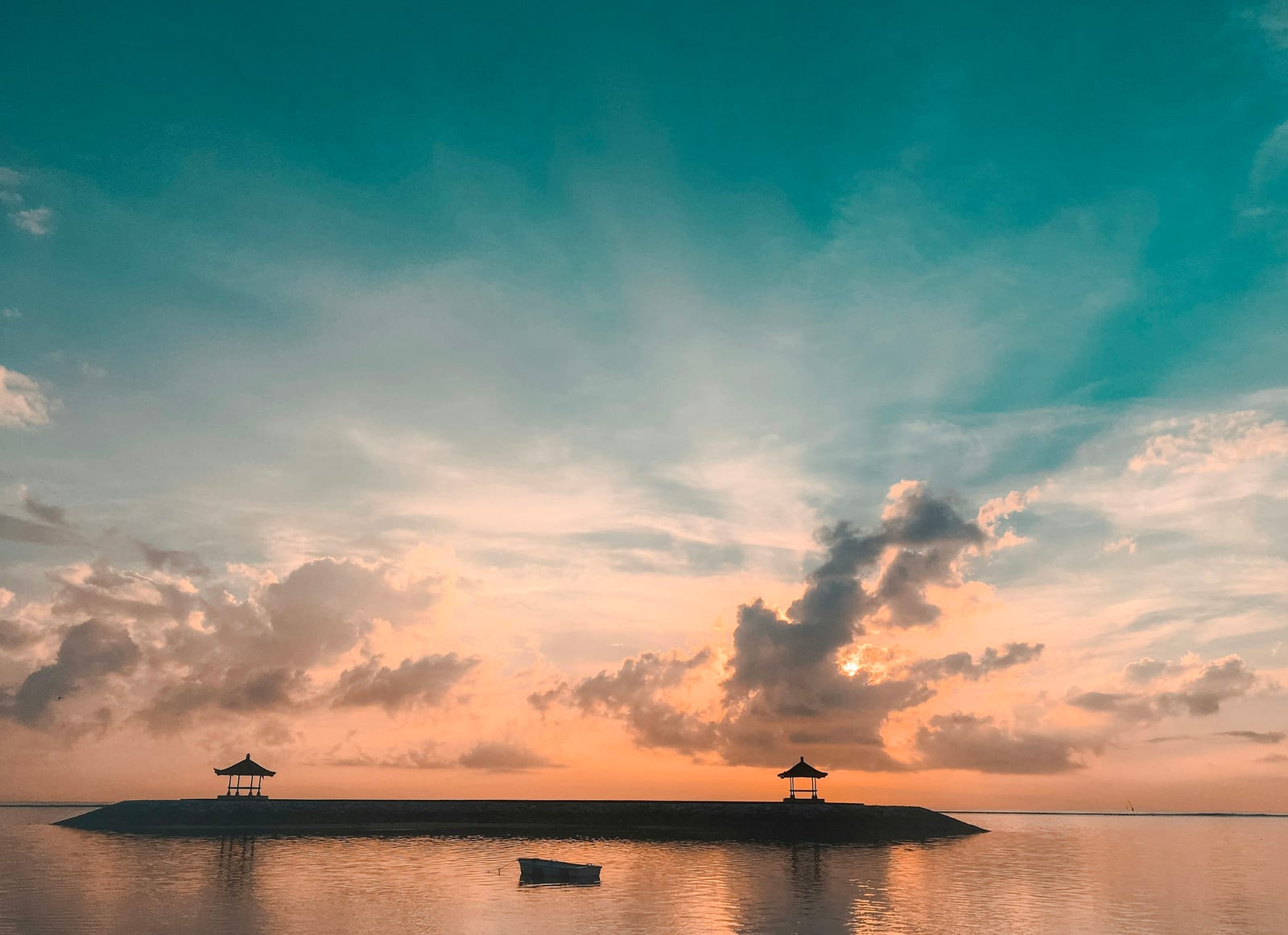 A bright sunrise colors the sky and calm sea behind two small shore pavilions at Sanur Beach.