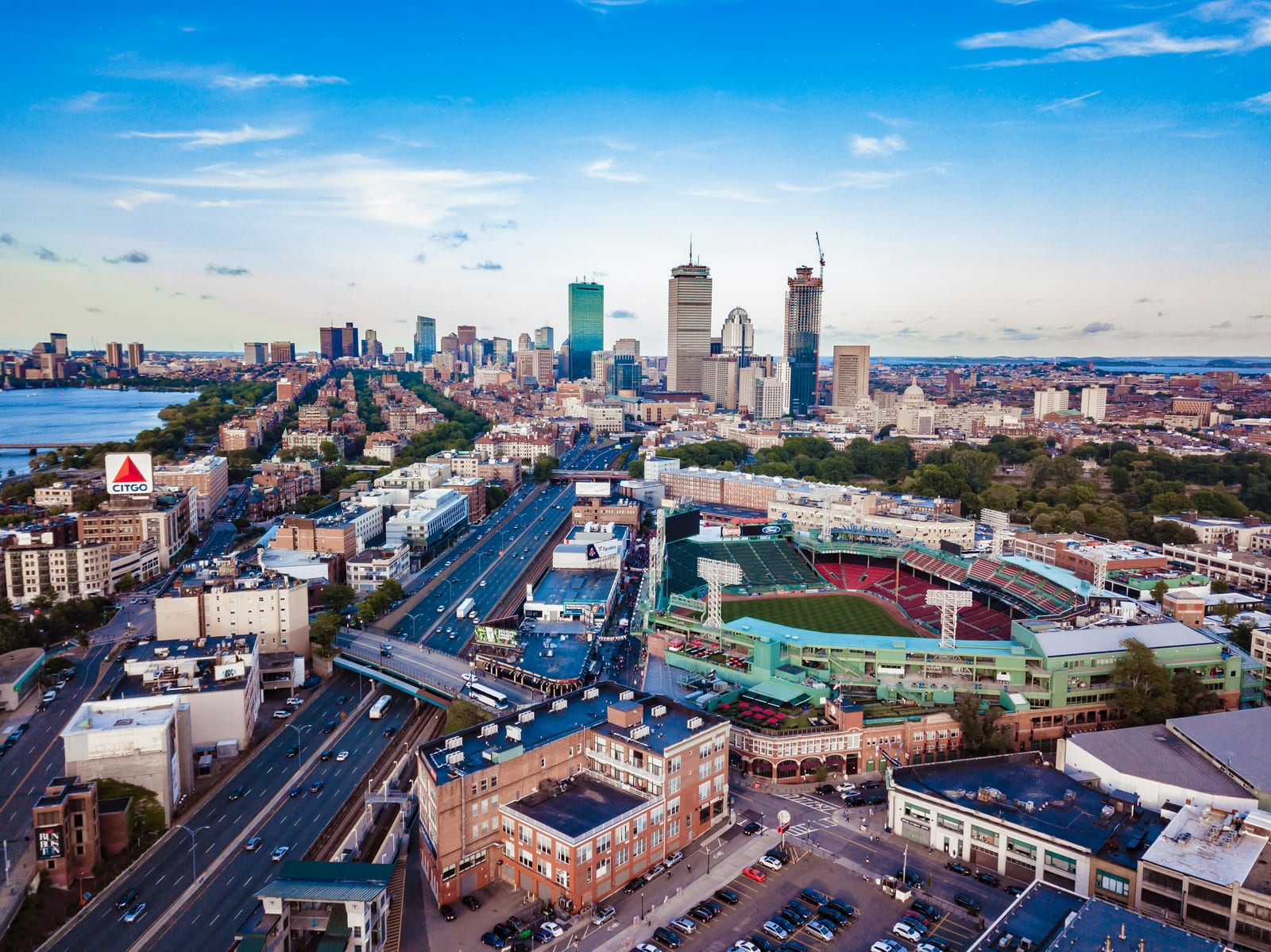 Aerial daytime view of Boston's skyline with Fenway Park and surrounding city streets visible in the foreground.