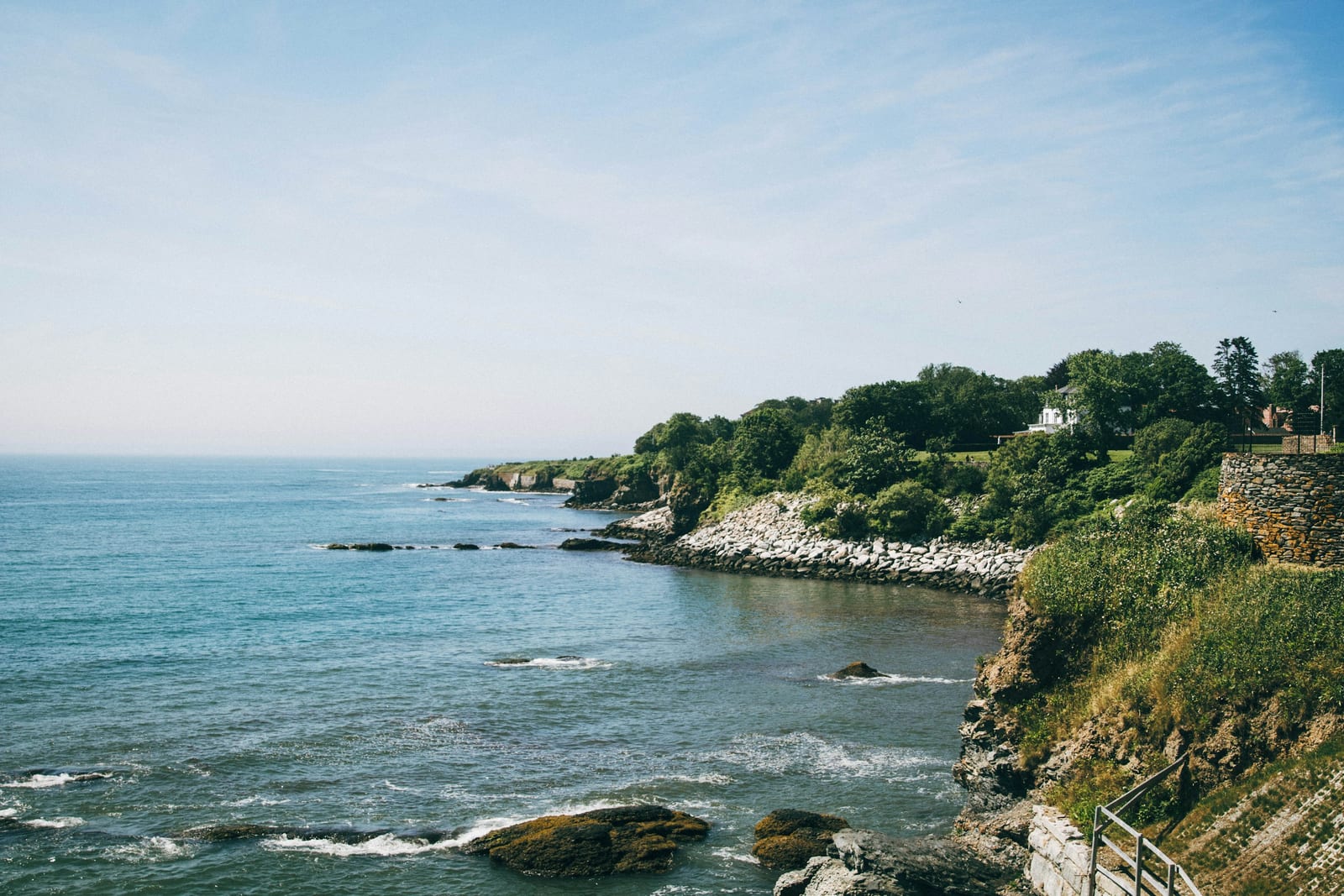 A rocky, tree-lined peninsula juts into calm blue water under a clear sky.