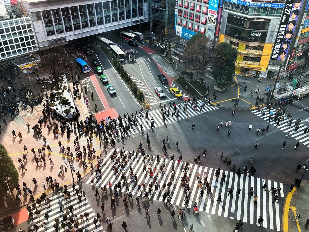 Crowds stream across the Shibuya scramble crossing seen from above on a bright day, ringed by billboards.