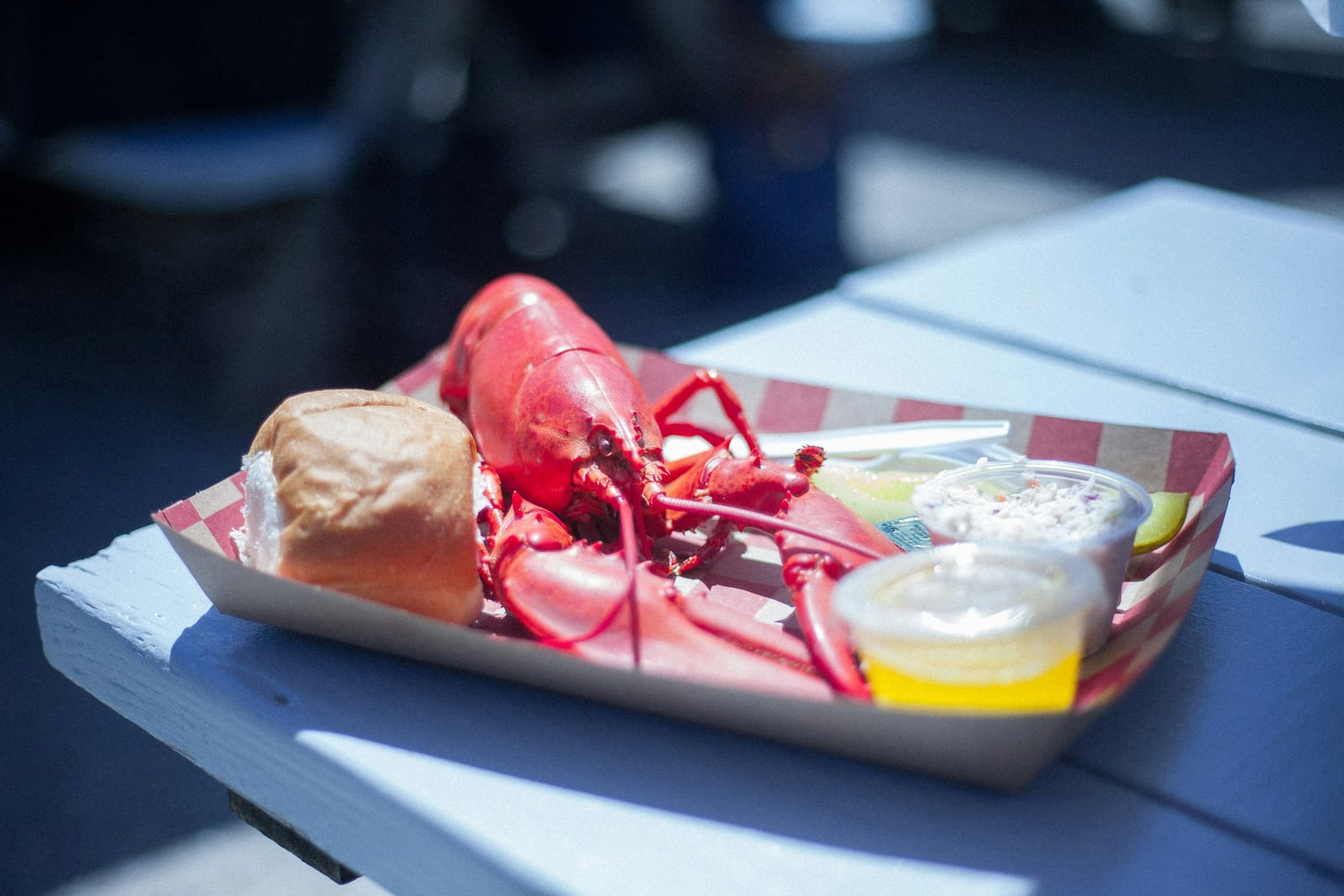 A whole steamed lobster with a buttered roll and a side of melted butter served in a red-and-white paper tray on a sunlit picnic table.