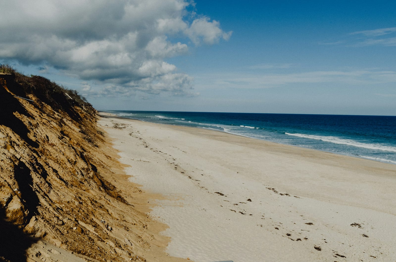 Eroded sand bluffs rise beside a wide, empty beach where blue Atlantic surf rolls in under a partly cloudy sky.