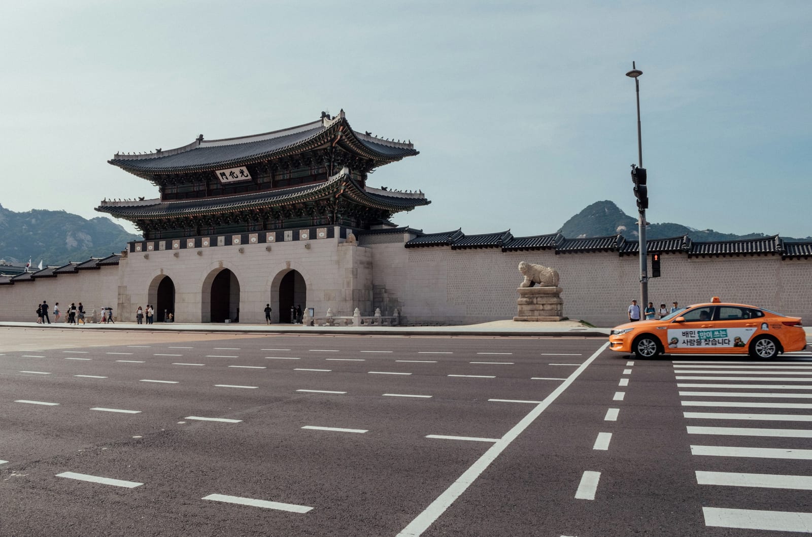 A Gyeongbokgung Palace gate stands beside a broad crosswalk, with a Seoul taxi and mountains behind it.