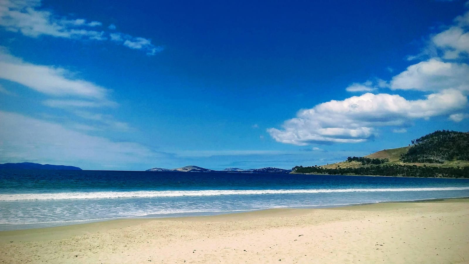 A wide sandy beach meets blue ocean waves beneath a bright blue sky with scattered clouds.