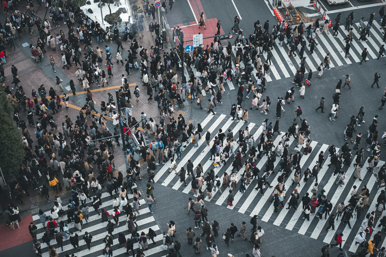 An aerial view of the Shibuya scramble crossing, with crowds streaming across its multi-directional crosswalks at dusk.