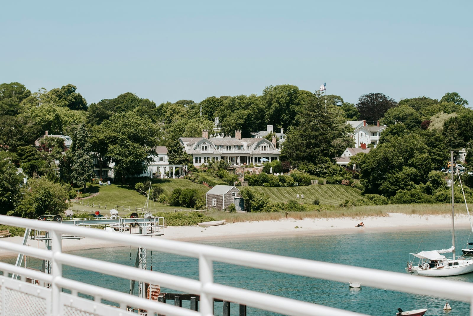 A sandy waterfront beach, sailboats, and large houses along a wooded shoreline.