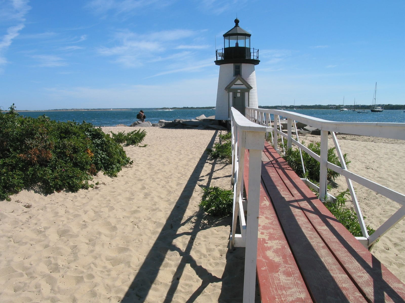 A white lighthouse stands at the end of a wooden boardwalk leading to a sandy beach under a clear blue sky.