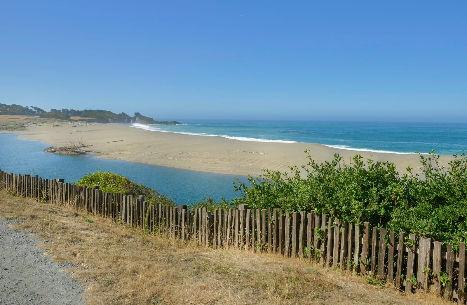 A sandy beach and inlet are viewed from a path behind a low wooden fence.