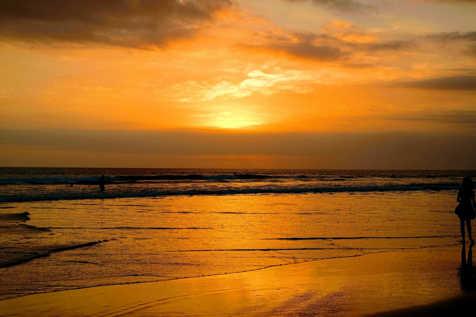 A person stands on a beach as the sun sets over the ocean, reflecting across the wet sand.
