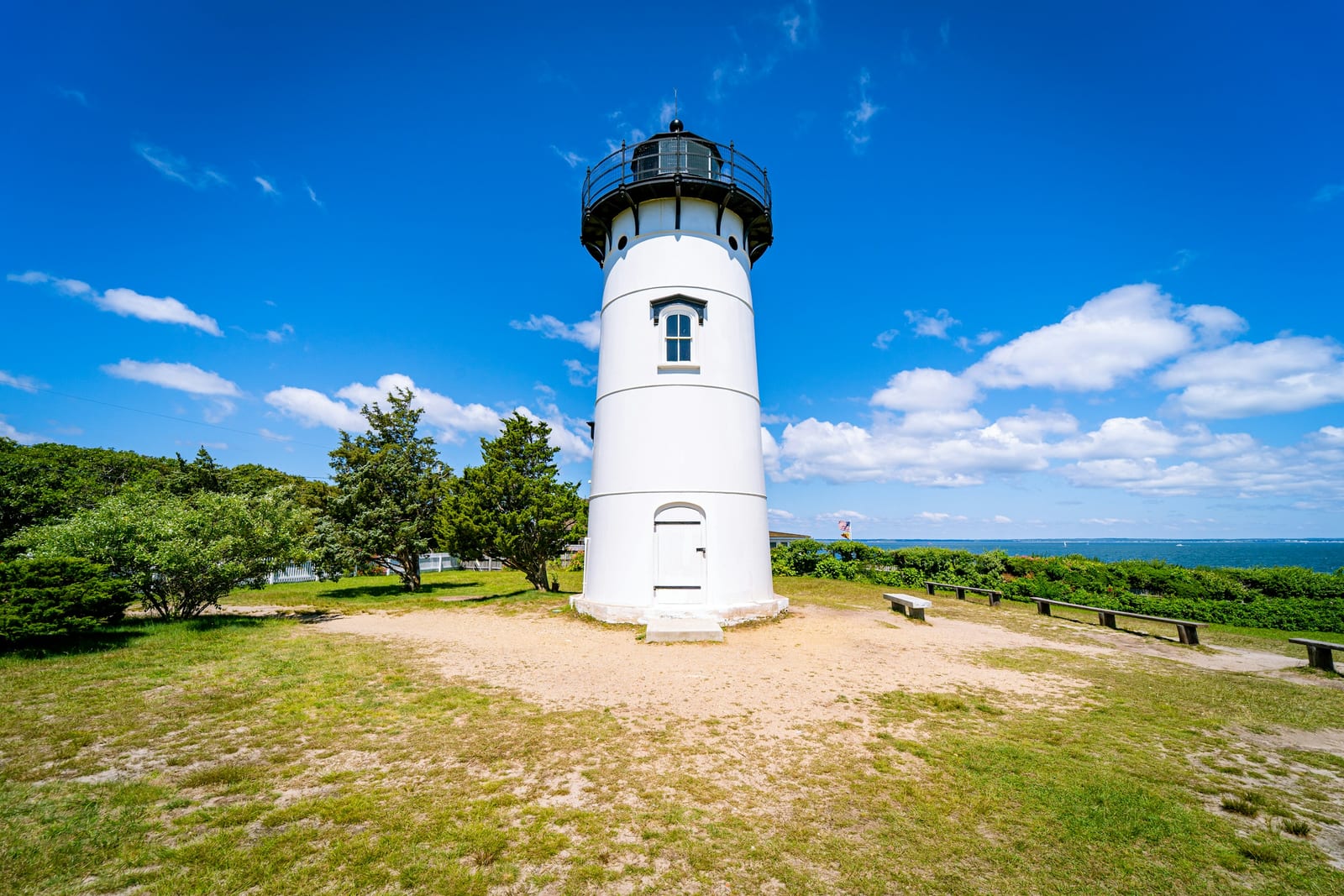 A white lighthouse stands on a grassy clearing with trees and blue ocean in the distance.