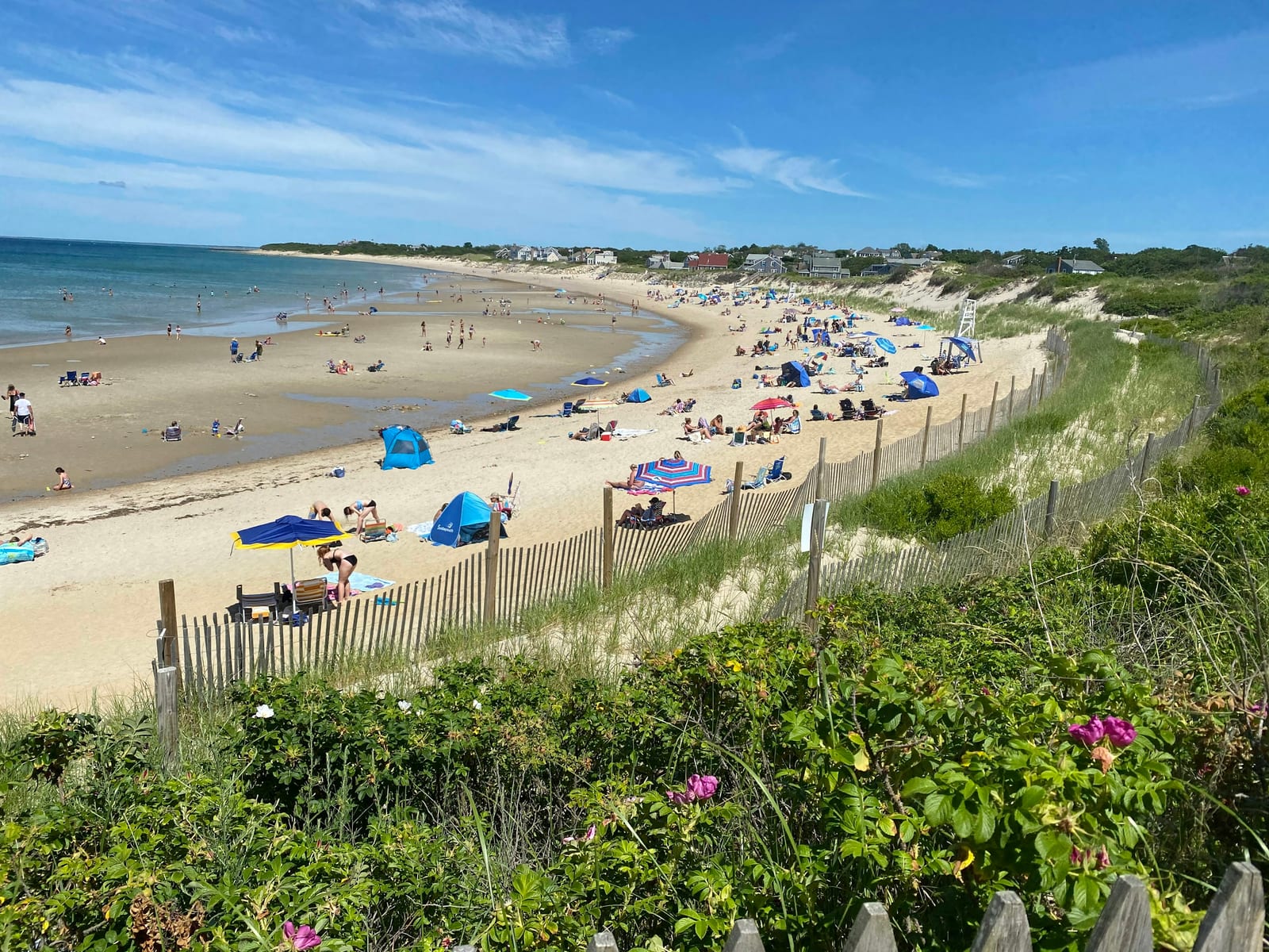 Crowded sandy beach with colorful umbrellas along a curving shoreline under a bright blue sky with a wooden fence in the foreground.