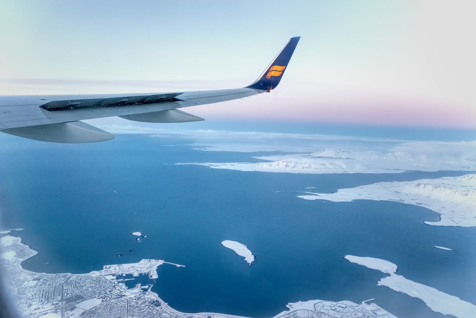An airplane wing extends over blue water, snowy coastline, and ice-covered land beneath a pale blue sky.