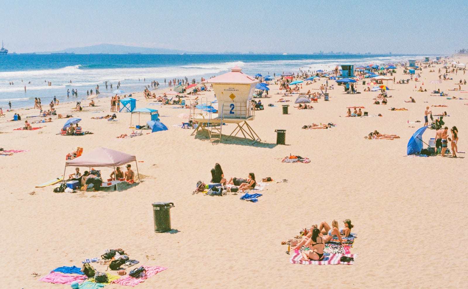 A bright public beach stretches along the ocean with swimmers, sunbathers, umbrellas, and a lifeguard station.