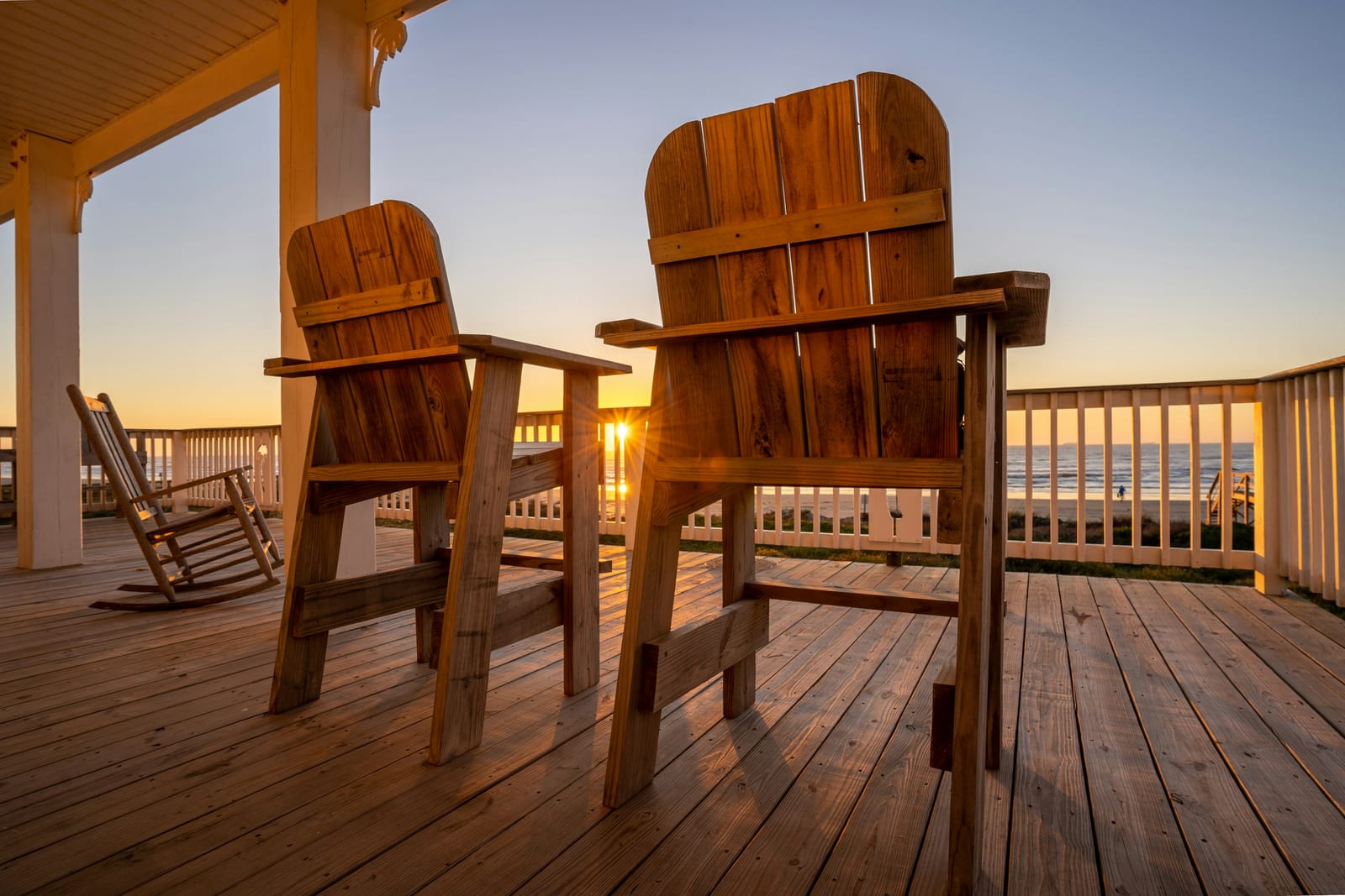 A sunlit beach house porch with two wooden Adirondack chairs, a rocking chair, and ocean views beyond the railing.