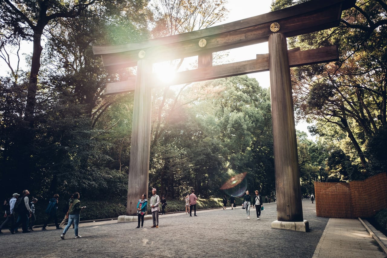 Visitors pass beneath the great wooden torii at the forest entrance to Meiji Jingu, the gravel approach lined with tall trees.