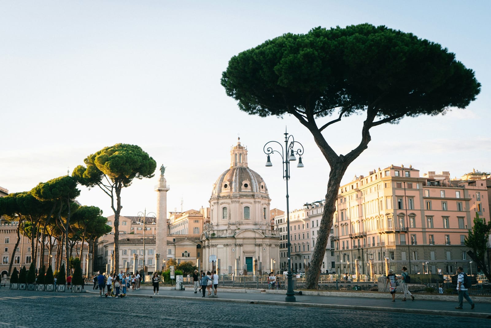 Golden evening light over Piazza Venezia in Rome, with umbrella pines and people strolling past the domes of Santa Maria di Loreto.
