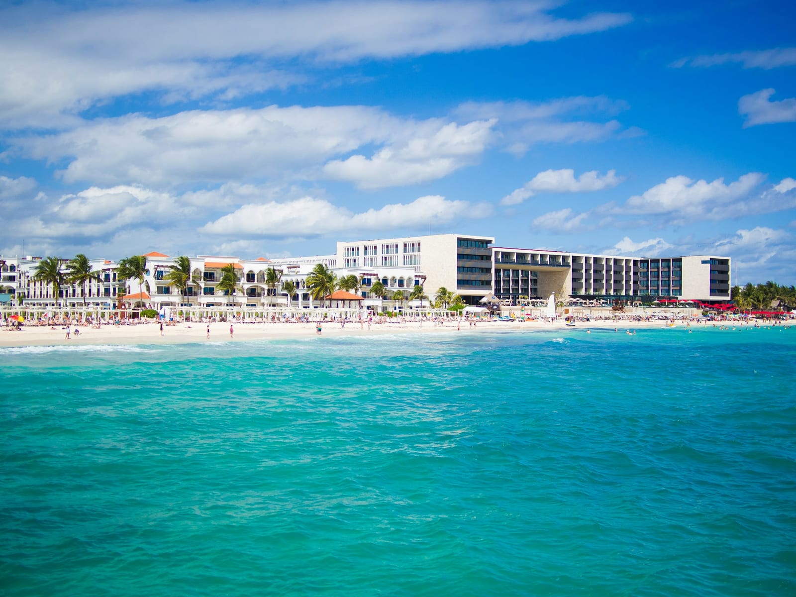 A beachfront hotel complex lines a sandy shore beside bright turquoise water under a blue sky.