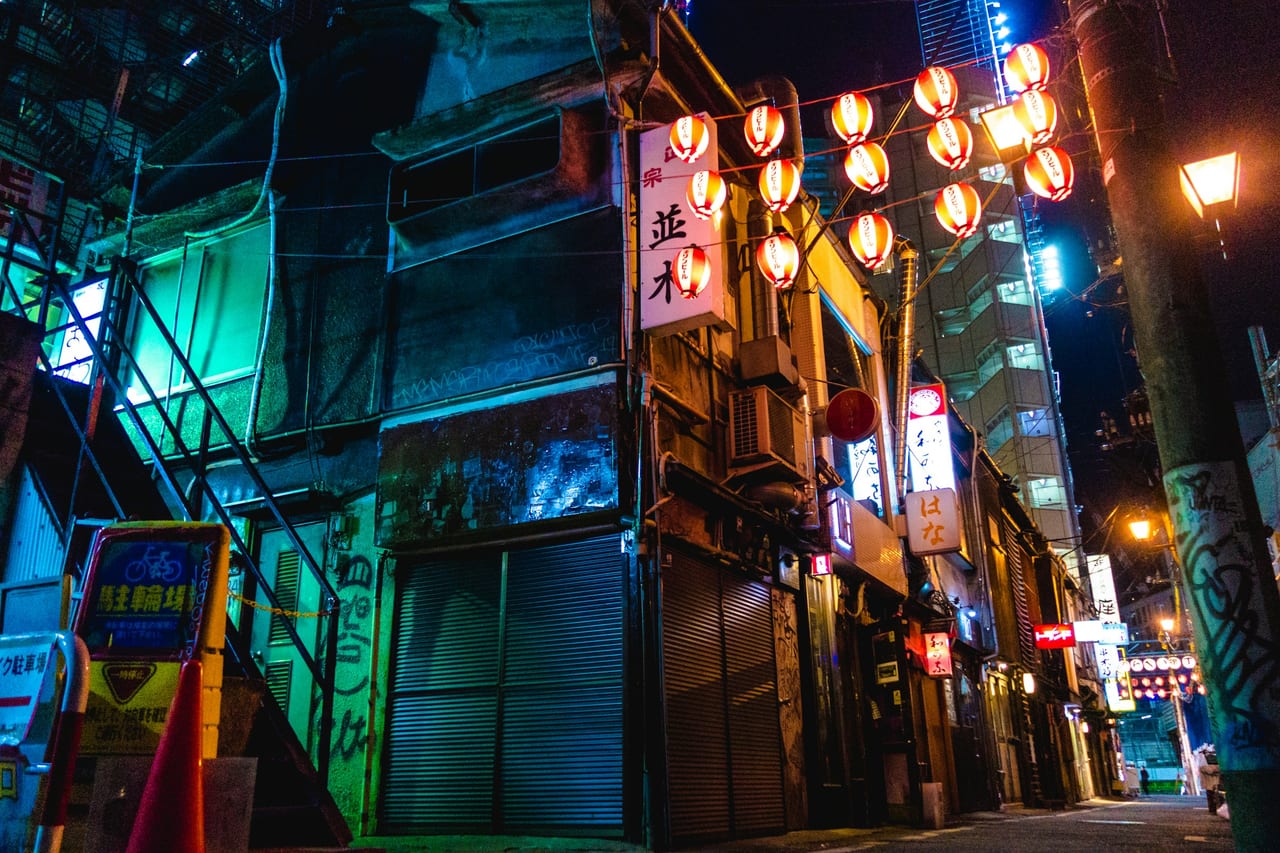 The tiny lantern-lit bars of Nonbei Yokocho line a narrow lane at night, with a modern Shibuya tower rising behind.