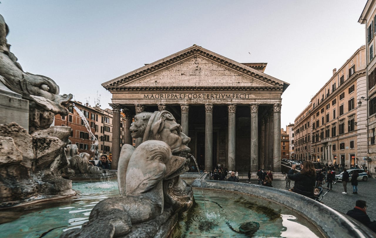 The Pantheon's columned portico and the fountain of Piazza della Rotonda under a bright sky.