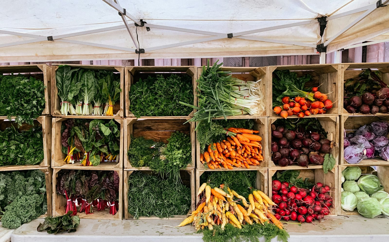 Wooden crates filled with fresh greens, carrots, and beets displayed at an outdoor market stall.