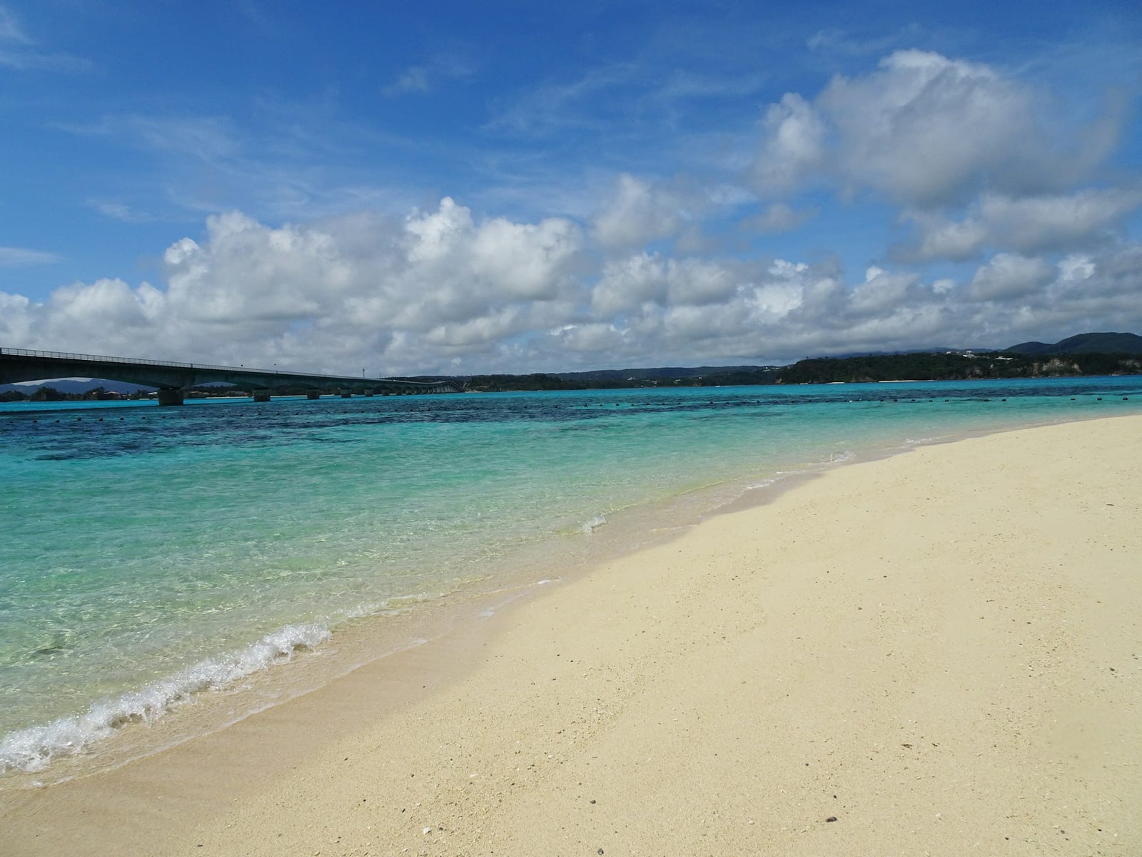A bright sandy beach meets clear turquoise water beneath a partly cloudy sky.