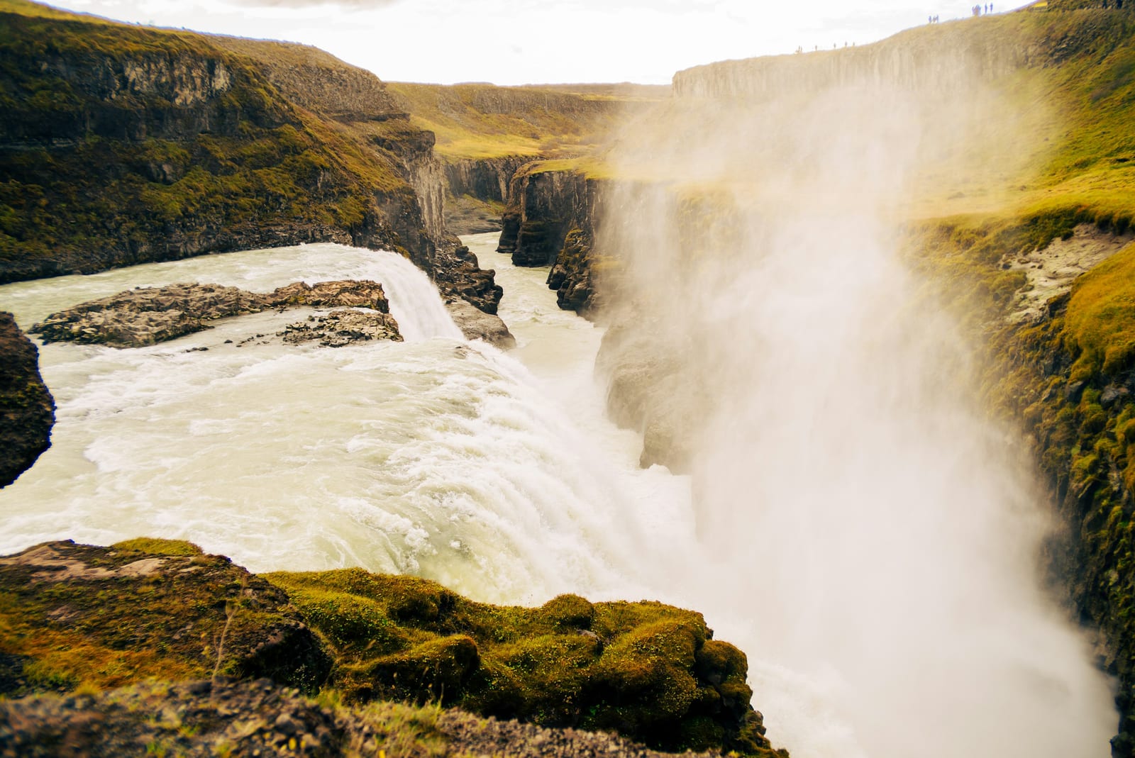 A broad Icelandic waterfall pours through a rocky gorge surrounded by green and golden hills.