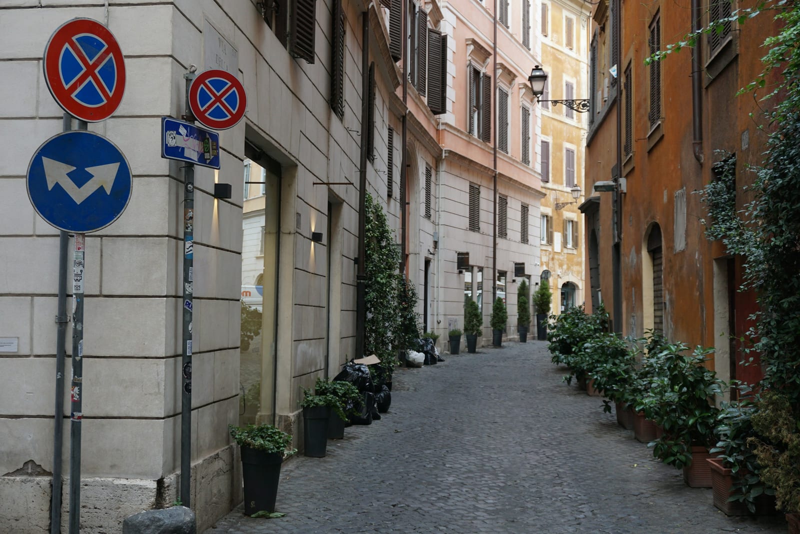 A quiet residential lane in Rome with ochre buildings, green shutters, potted plants, and cobblestones.