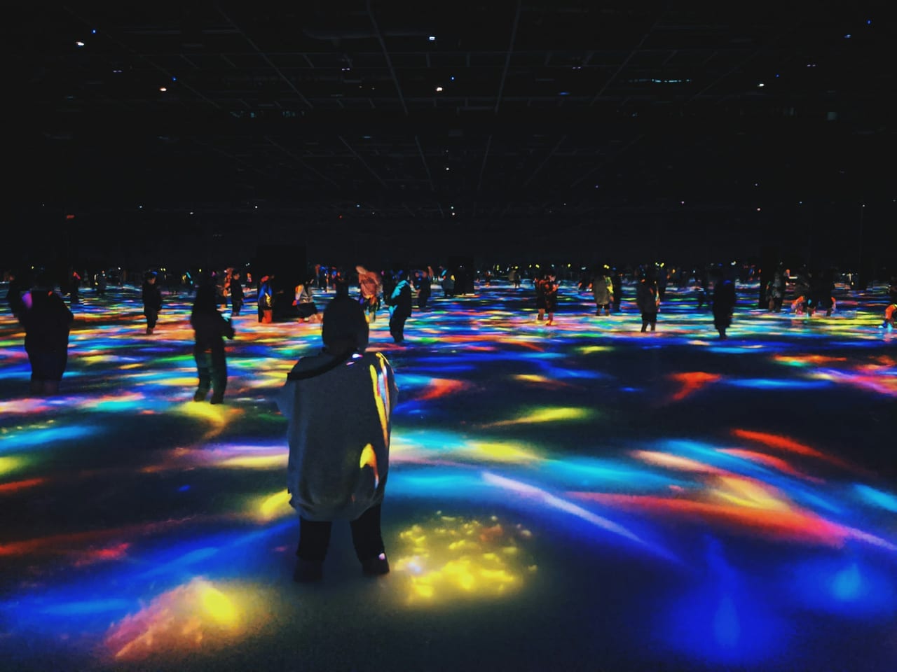 Visitors wade through a darkened teamLab room as projected light blooms across the walls and water.