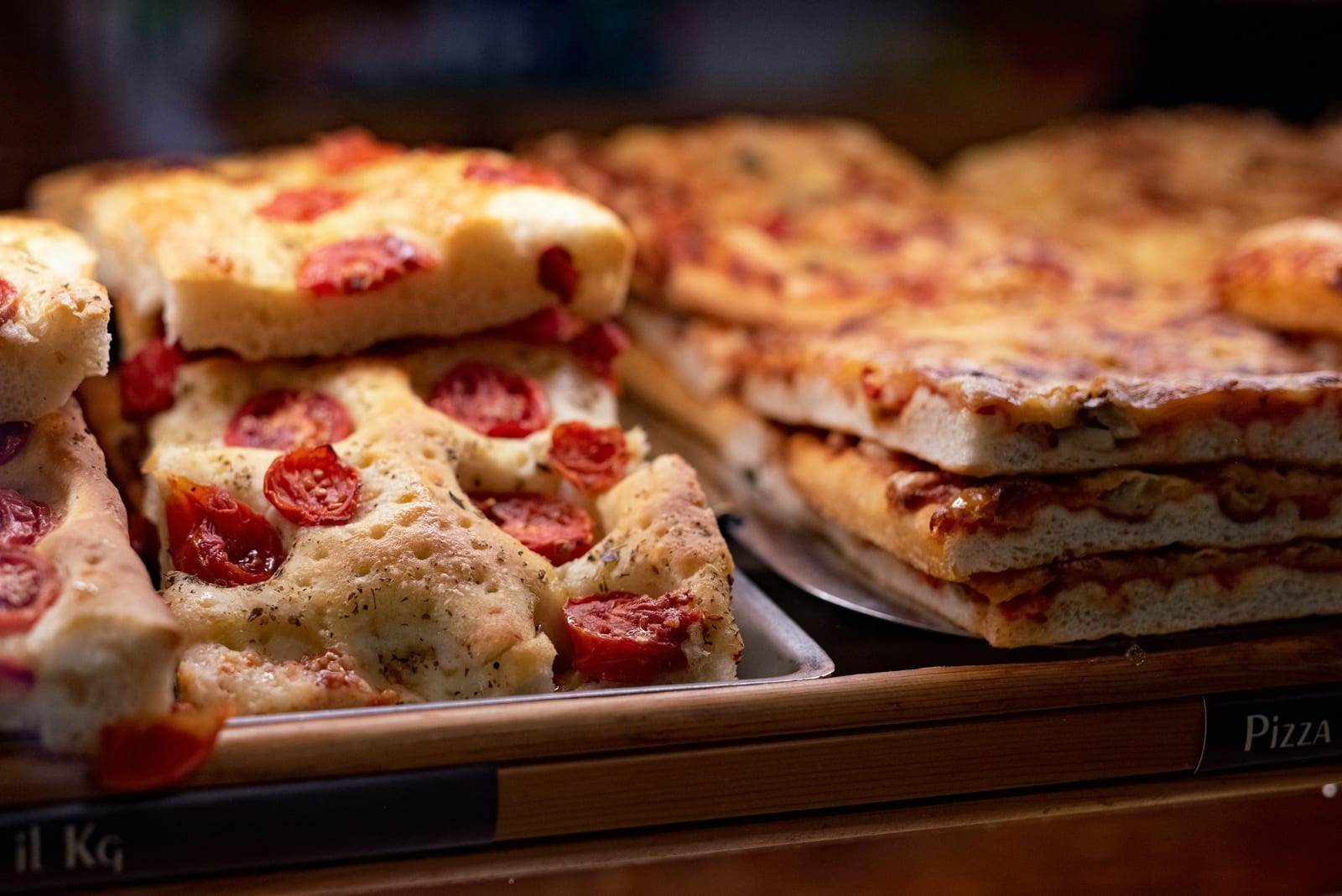A display counter of Roman pizza al taglio topped with tomatoes, greens, and vegetables, sold by the slice.