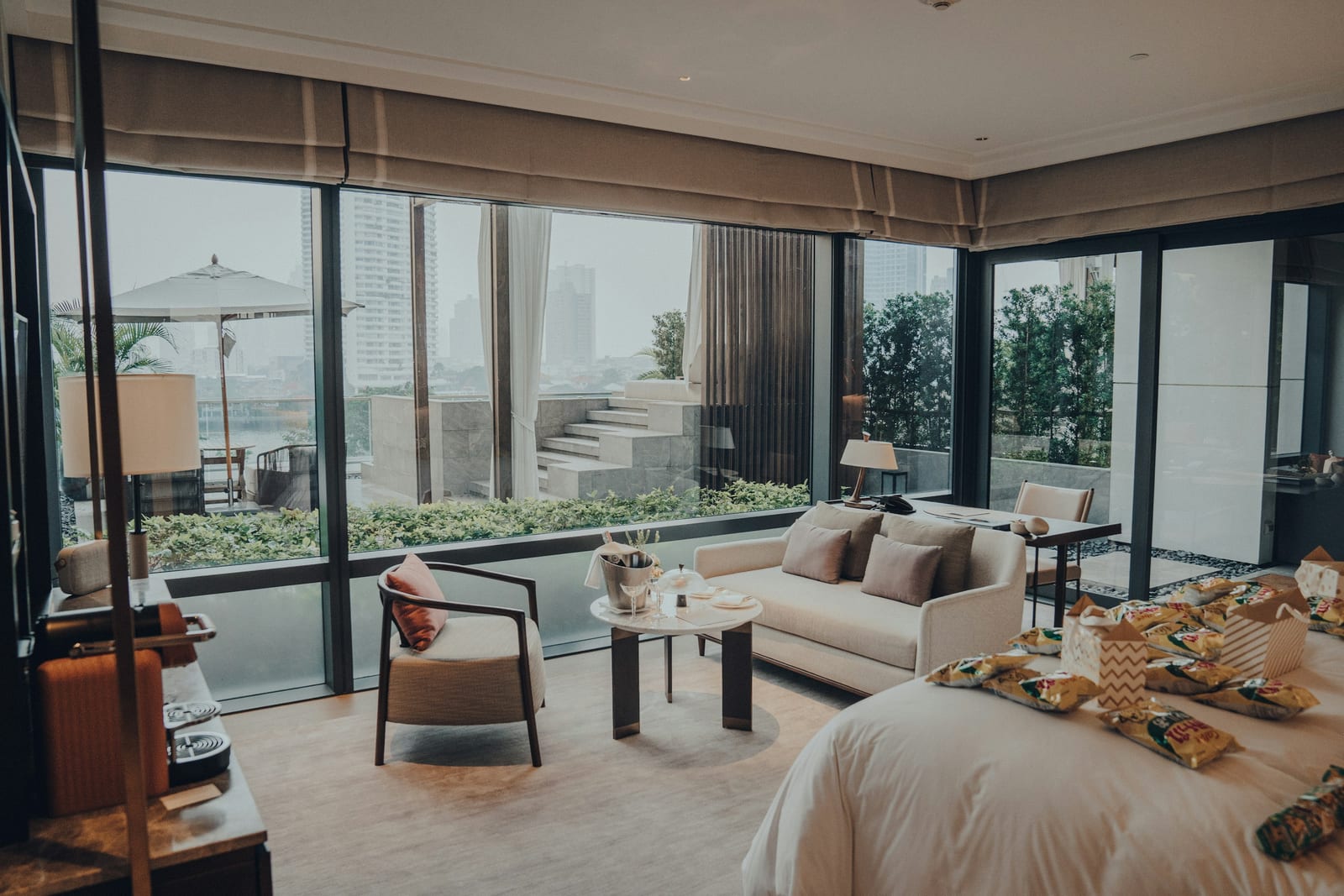 A stylish hotel room with a bed, seating area, dining table, and floor-to-ceiling windows overlooking a city view.