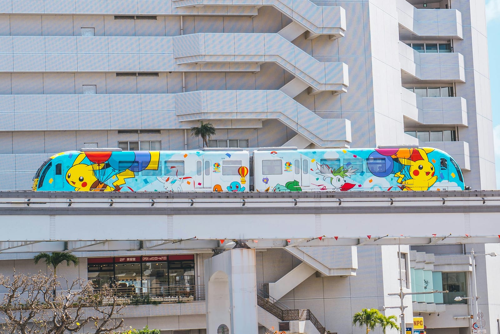 A colorful monorail passes along an elevated track beside city buildings and palm trees.